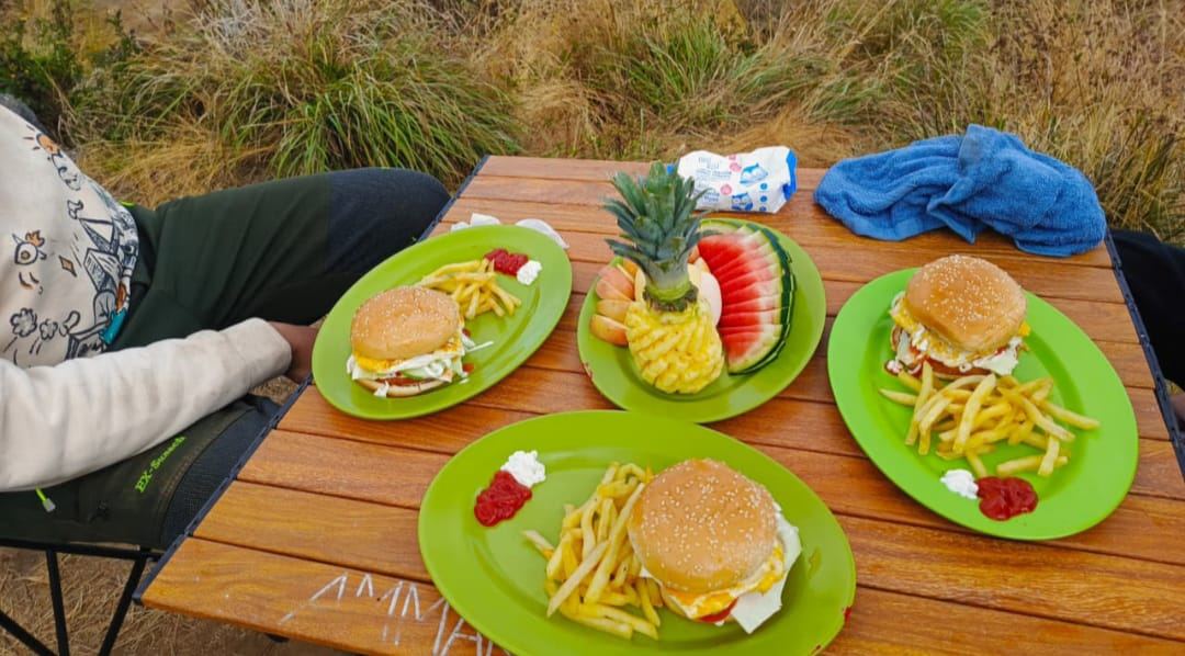 Four green plates on a camping table on the mountain: burgers with fries and ketchup, and a carved pineapple with watermelon and fresh fruit