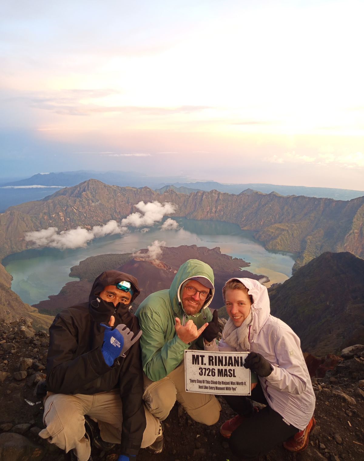 Three trekkers at the Mt Rinjani summit sign at sunrise, with the crater lake far below them